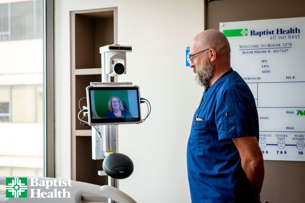 A Baptist Health bedside care team member welcomes a virtual clinician into the patient room using Caregility’s APS250C mobile telehealth cart.